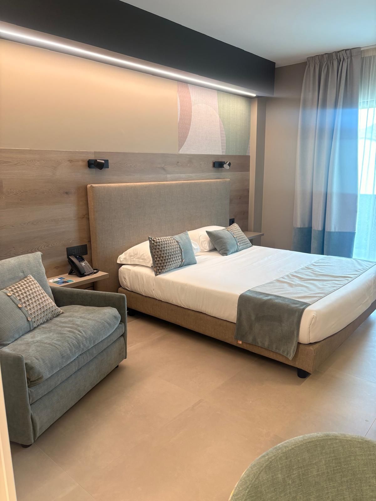A modern hotel room with a large bed, a sofa, a bedside table with a phone, and a wall-mounted artwork.