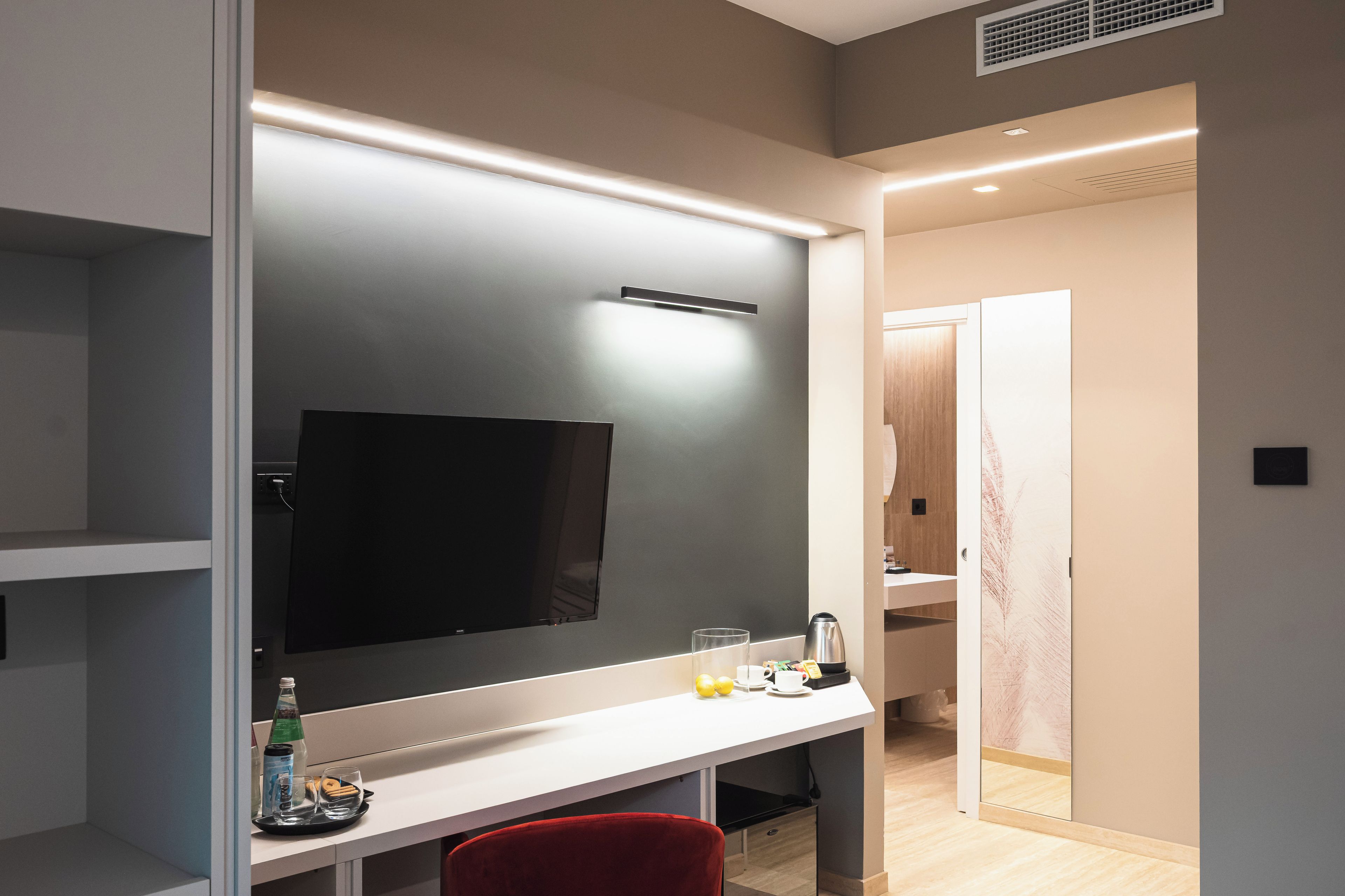 A modern hotel room with a large flat-screen TV, a desk with a tray of breakfast items, and a red chair.