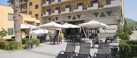 A hotel patio with outdoor seating and umbrellas, a palm tree, and a multi-story building in the background.