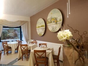 Comedor con mesas dispuestas para cuatro, un ventanal con cortinas y cuadros con paisajes.