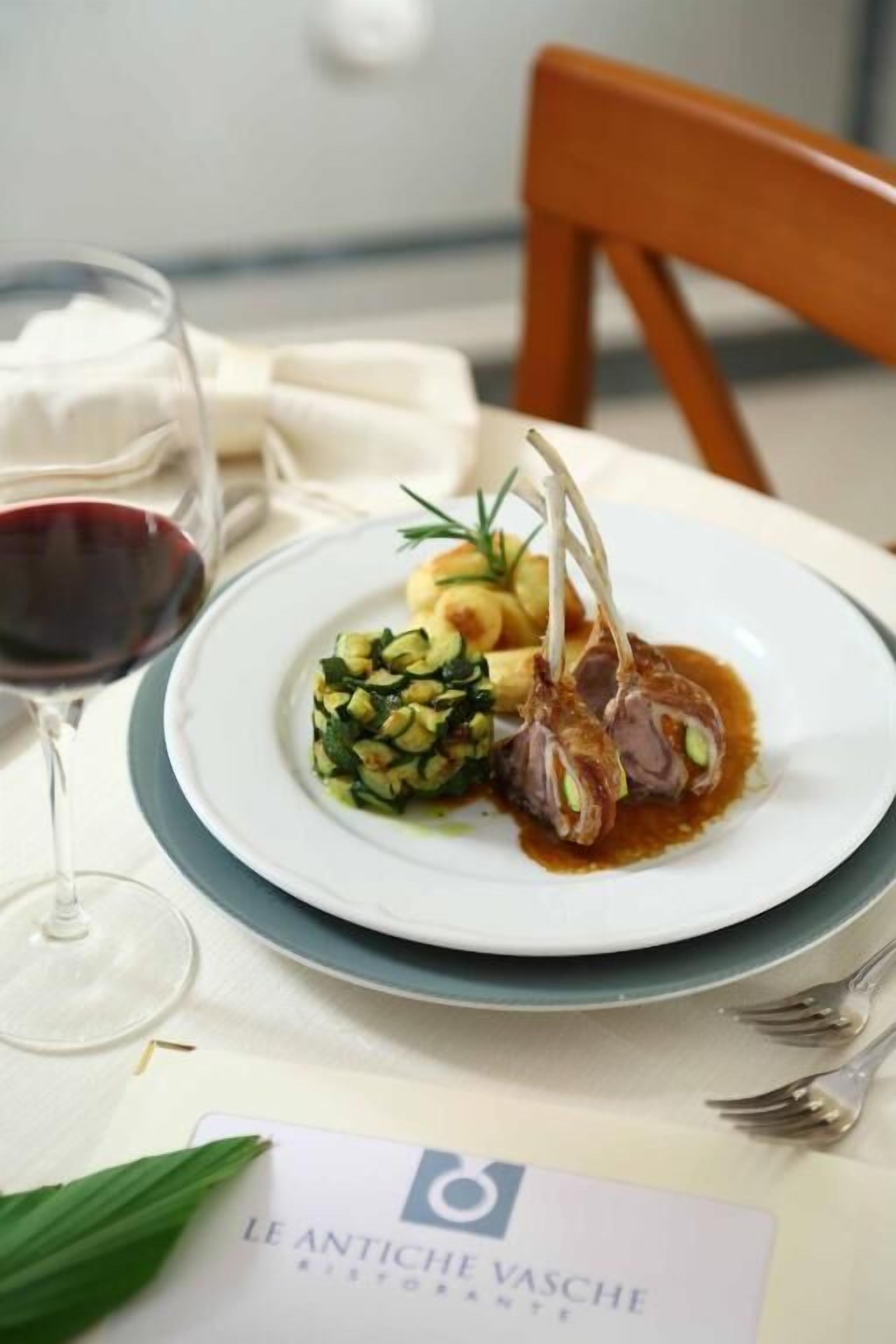A plate of food with lamb chops, a side of vegetables, and a glass of red wine on a table with a placemat and a chair.