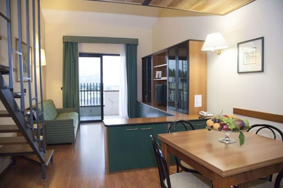 Kompakti hotellihuone, jossa on pieni keittiönurkkaus, ruokailutila hedelmäkulhon kanssa, portaikko ja ikkunasta avautuva näkymä, jossa on verhot.