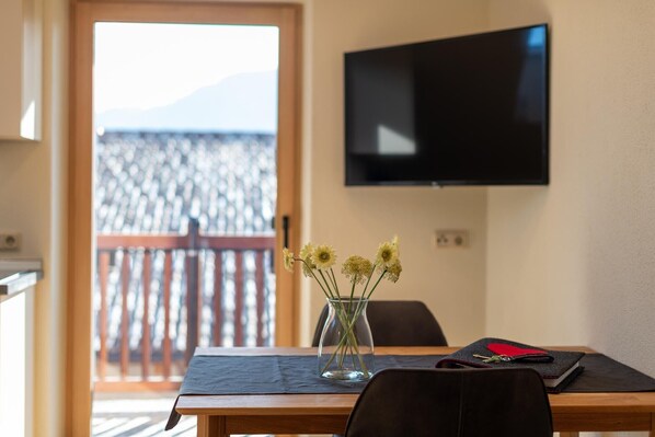 A dining area with a table, chairs, a vase with flowers, a television on the wall, and a view of a balcony.