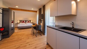 Una habitación de hotel moderna con suelo de madera, cama con ropa de cama roja y blanca, un pequeño comedor y una cocina con un fregadero y armarios.