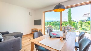 A modern living room with a wooden dining table, black leather chairs, a large window with a view of greenery, and a television on the wall.