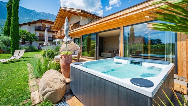 A modern outdoor hot tub with a wooden deck, a glass-enclosed patio, and a mountain backdrop.