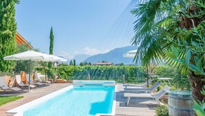 A swimming pool with lounge chairs and umbrellas, surrounded by greenery and mountains in the background.