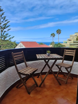Una terraza con dos sillas de madera y una mesa, una planta en maceta, y vistas al océano y edificios.