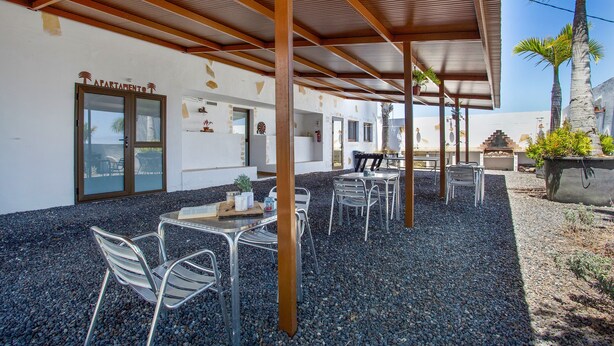 Un patio al aire libre con sillas y mesas de metal, una zona de estar cubierta y la entrada de un edificio con la leyenda 'APARTMENT'.