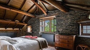 A rustic room with a stone wall, wooden ceiling beams, a bed with a white bedspread, a wooden dresser, and a window with a view of trees. - Holiday Home 'Mirador Del Navia' with Mountain View, Private Terrace and Wi-Fi (Serandinas)