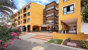 A multi-story yellow building with balconies, a covered entrance, and a small garden area. - Vacation Apartment 'Apto El Mocan Swimming Pool 2bed' with Shared Pool, Private Terrace and Wi-Fi (Palm-Mar)