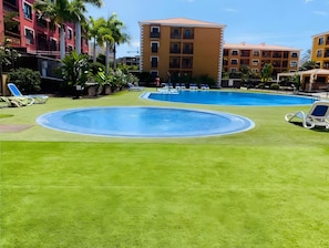 Piscina com área gramada ao redor, cadeiras de espreguiçadeira e um edifício de vários andares ao fundo.