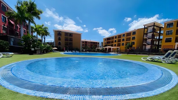 A swimming pool with a tiled edge, surrounded by grass and palm trees, with multi-story buildings in the background. - Vacation Apartment 'Apto El Mocan Swimming Pool 2bed' with Shared Pool, Private Terrace and Wi-Fi (Palm-Mar)