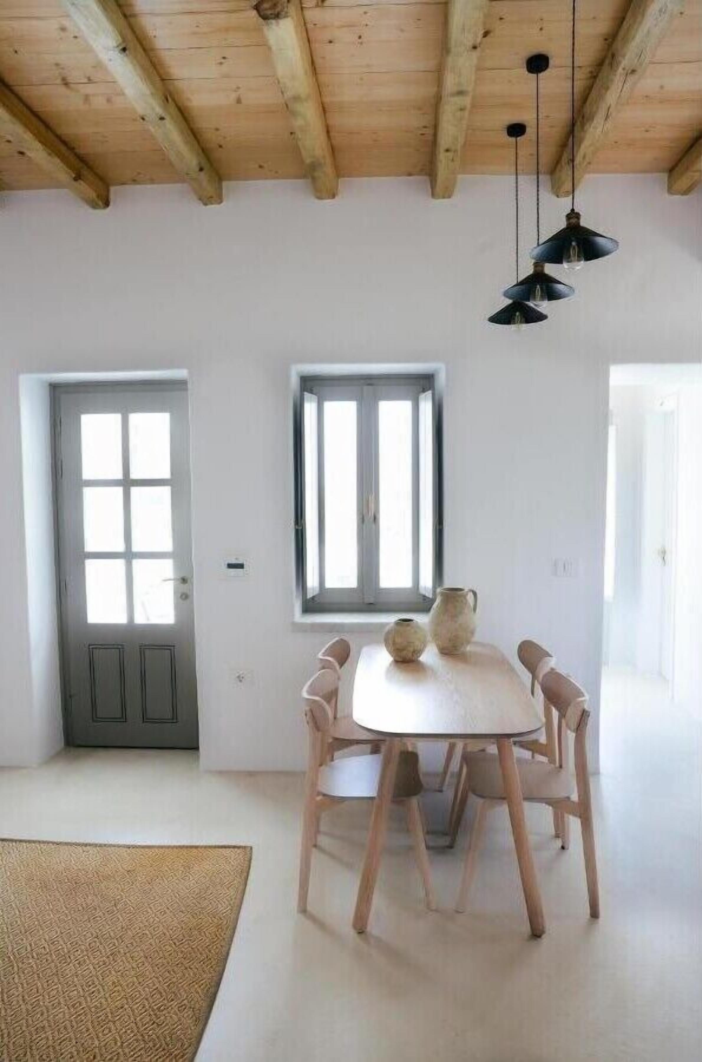 Una habitación con techo de madera, una mesa blanca con sillas, una ventana y una puerta.