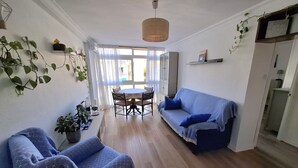 A living room with a sofa, a dining table with chairs, a hanging light fixture, and a window with curtains.