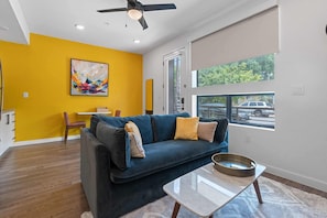 A modern living room with a dark blue sofa, a glass coffee table, and a painting on the wall. - Cozy DT ATX Studio | Mins to Rainey St | Patio (Austin)