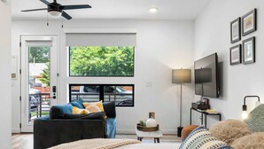 A modern hotel room with a bed, a sofa, a TV, a ceiling fan, and a large window with a view of greenery. - Charming DT ATX Studio / Mins to Rainey St / Patio (Austin)