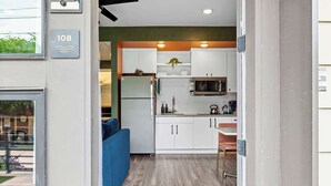 Una cucina moderna con mobili bianchi, un forno a microonde e un frigorifero. Si nota un divano blu e un tavolo da pranzo con sedie.