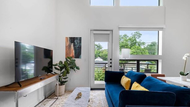 A modern living room with a flat-screen TV, a blue sofa, a coffee table, and a large window offering a view of greenery. - Charming DT ATX Loft | Near Rainey St (Austin)