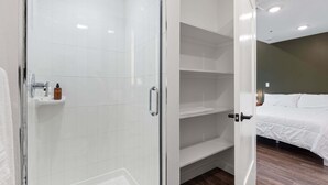 A modern bathroom with a glass-enclosed shower, a white sink with toiletries, and a bedroom with a bed and shelves.