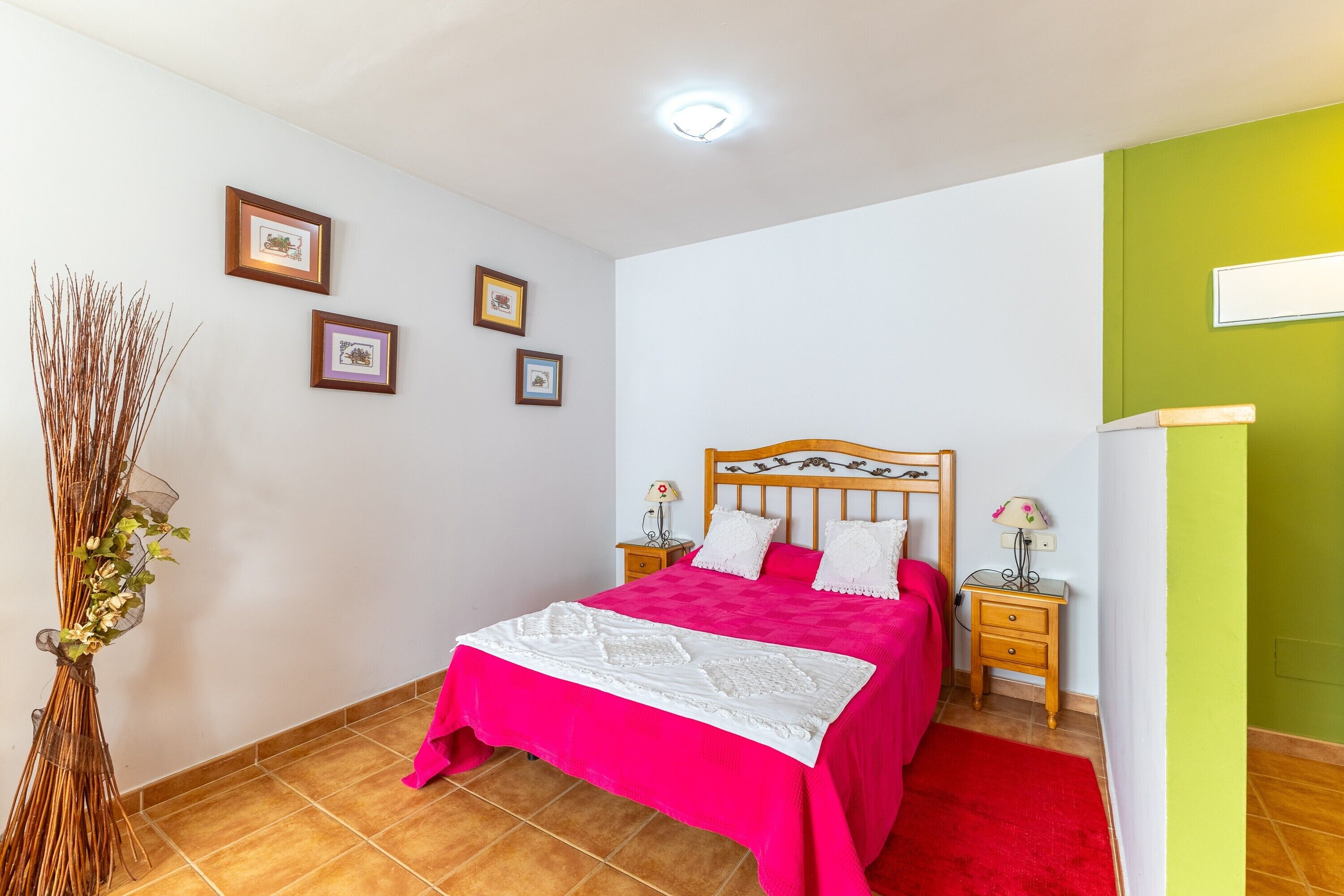 A bedroom with a bed, bedside tables, a nightstand, a wall-mounted lamp, and a vase with dried plants.