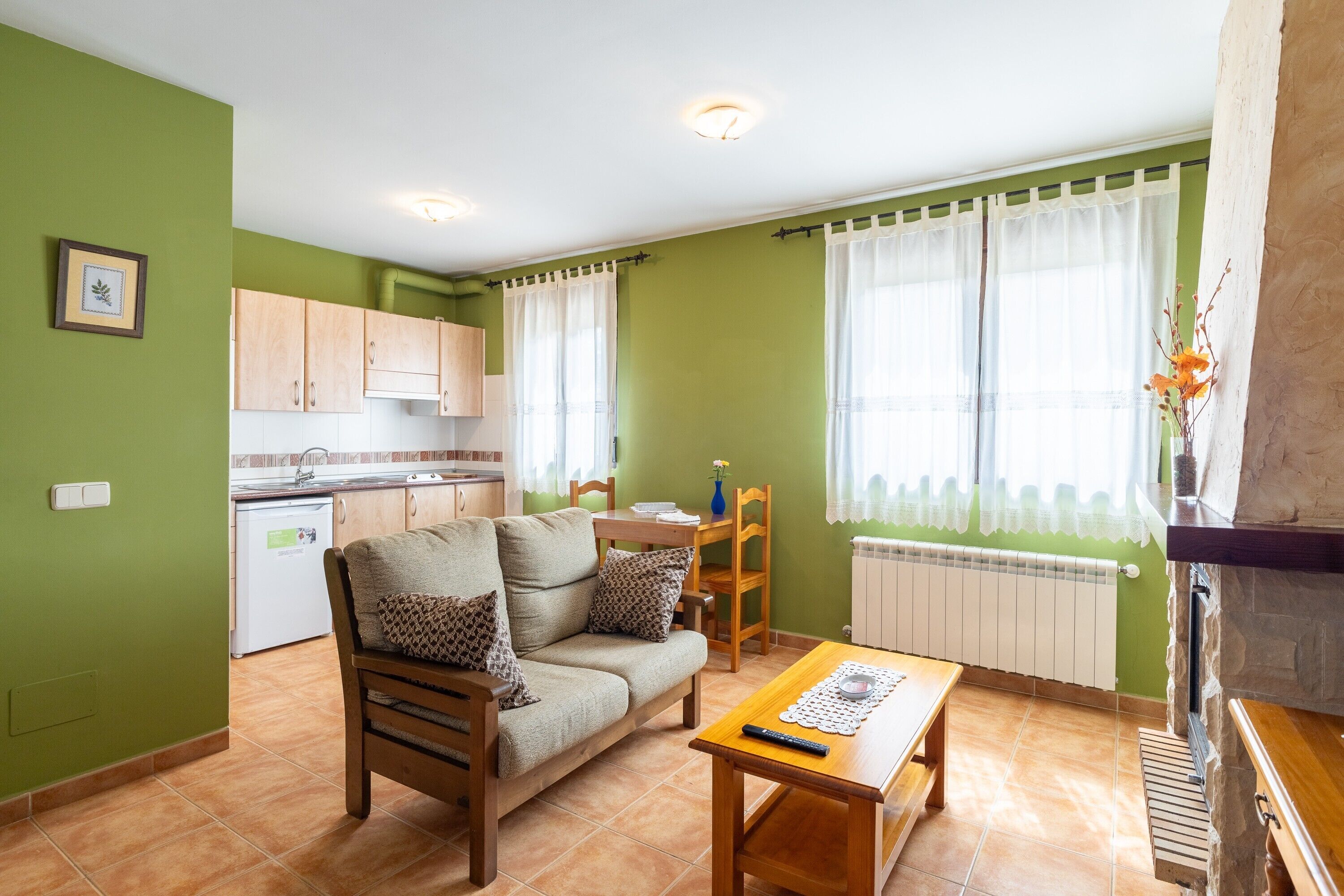 A living room with a sofa, a wooden table, a kitchen in the background, and a window with curtains.