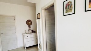 Ein Flur mit weißen Wänden, einer weißen Tür, einer weißen Kommode mit Schubladen und einem gerahmten Bild an der Wand.