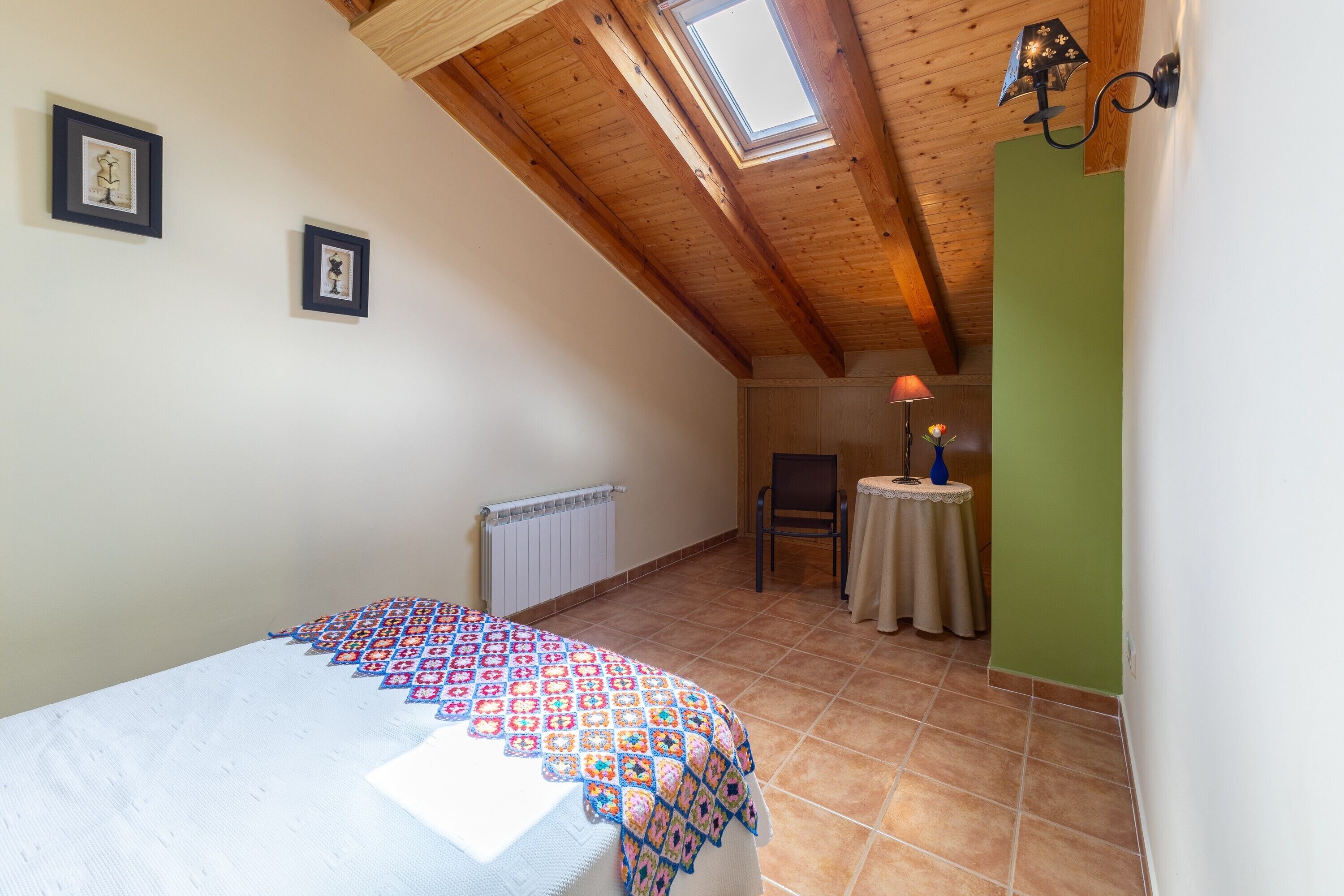Un dormitorio con techo inclinado, tragaluz, cama con manta estampada, radiador, silla y mesita de noche con lámpara.