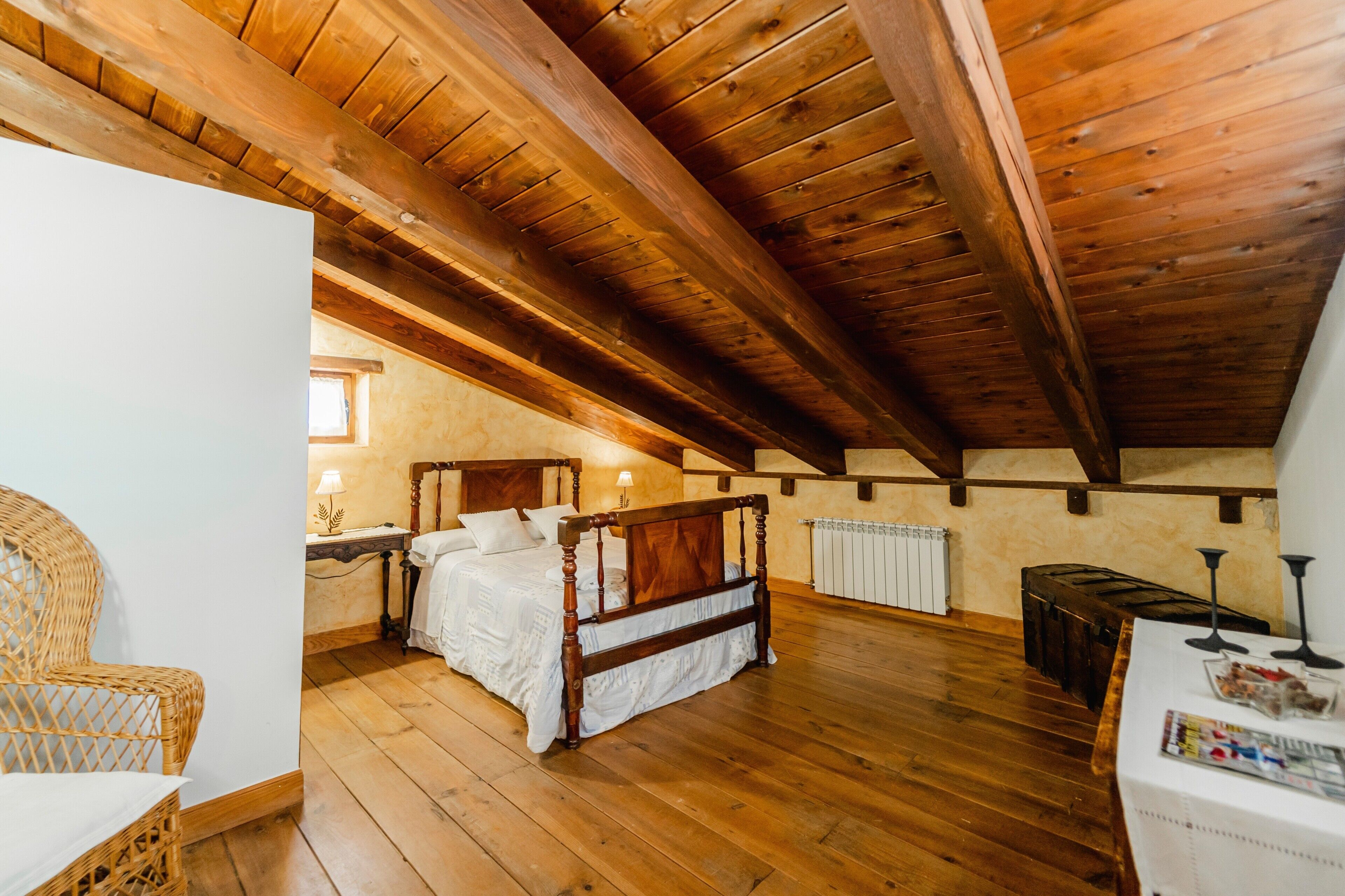 A bedroom with a wooden ceiling, a bed, a nightstand, a wicker chair, and a table with a magazine.