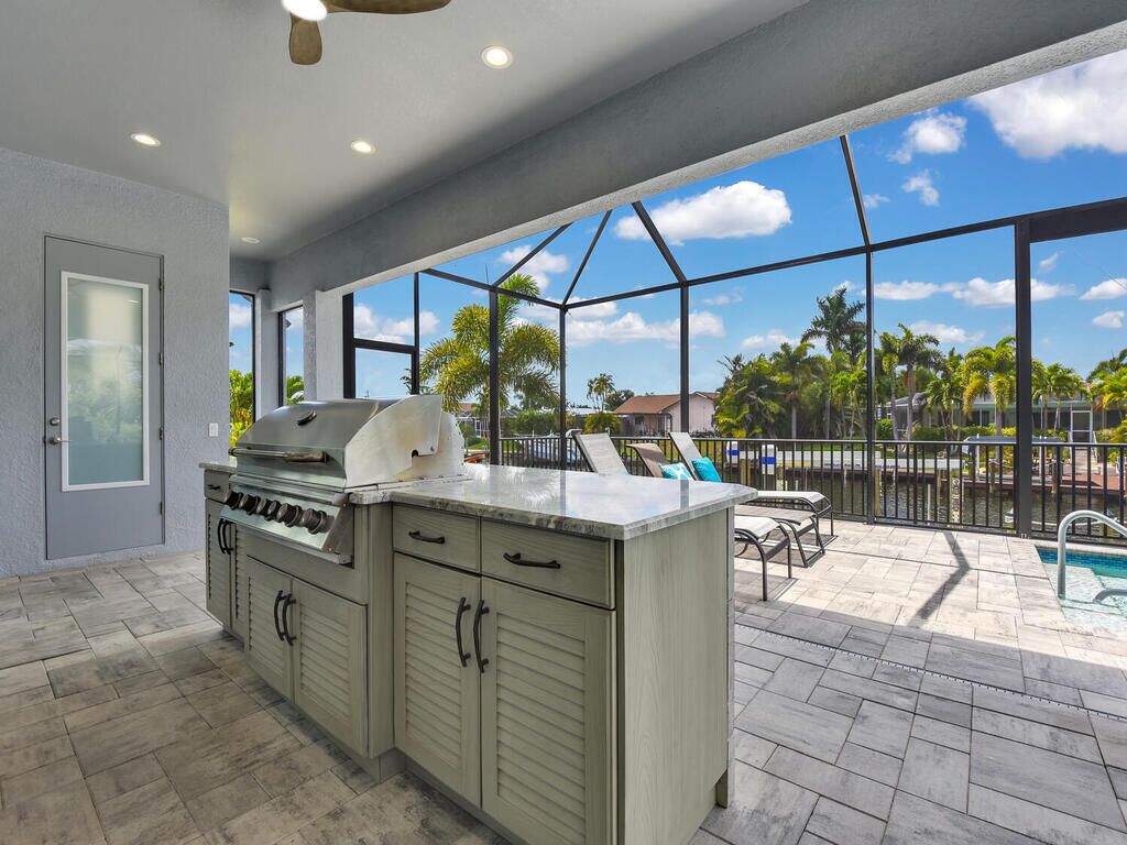 A kitchen with a grill, a dining area with chairs, and a view of a swimming pool and palm trees.
