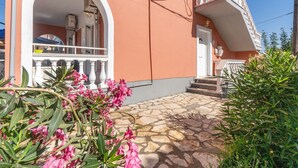 Un bâtiment rose avec un balcon blanc, des marches en pierre et un jardin où poussent des fleurs roses.