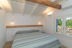 A bedroom with a wooden headboard, a bed with a striped bedspread, a window with a view of greenery, and recessed ceiling lighting. - Holiday Home 'Villa Cactus, Costa Corallina' with Sea View, Wi-Fi, and Air Conditioning (OLbia)
