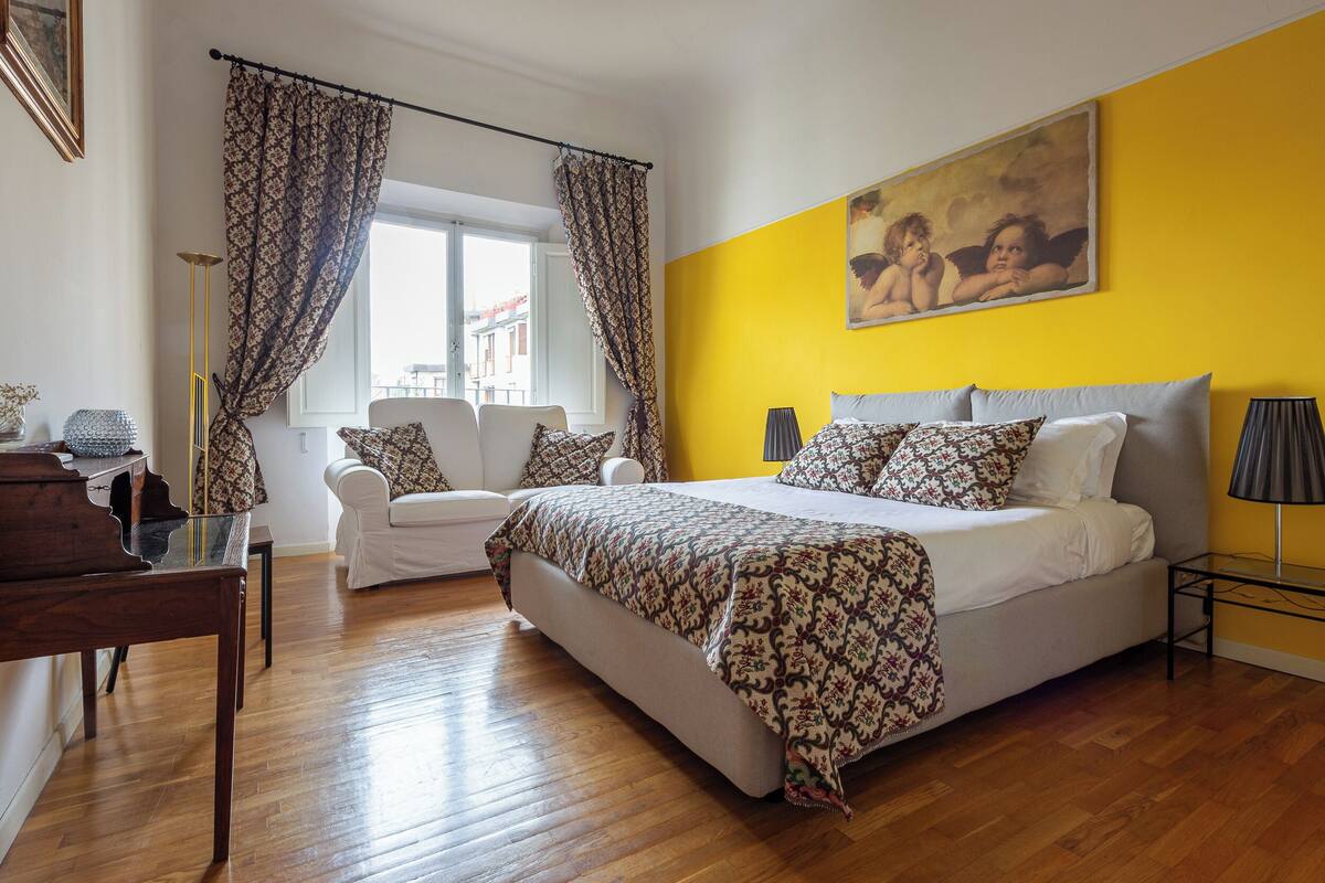 A bedroom with a large bed, a white sofa, a desk, and a window with curtains.