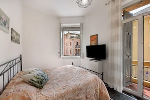 Quarto com cama, televisão, janela com vista para edifícios e porta para uma varanda.
