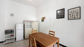 Una cocina pequeña con microondas, refrigerador y una mesa de comedor de madera con sillas. Hay luminarias fijadas a la pared e impresiones botánicas enmarcadas.