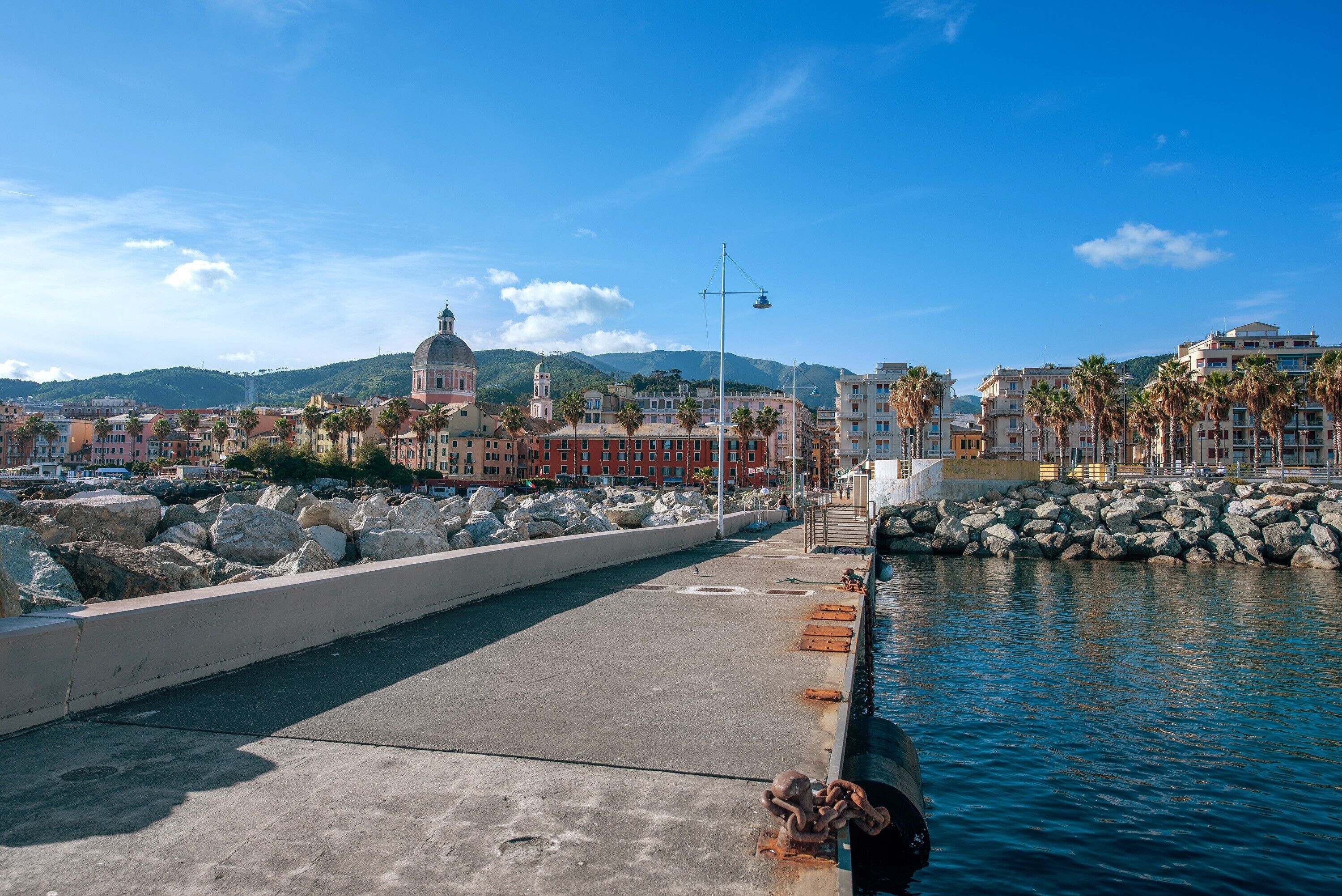 A waterfront promenade with a stone barrier, a clear sky, and buildings in the background.