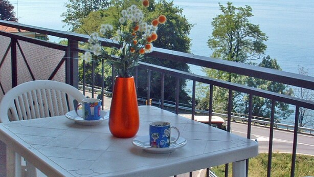 A balcony with a table, chairs, a vase with flowers, and a view of a lake and mountains.