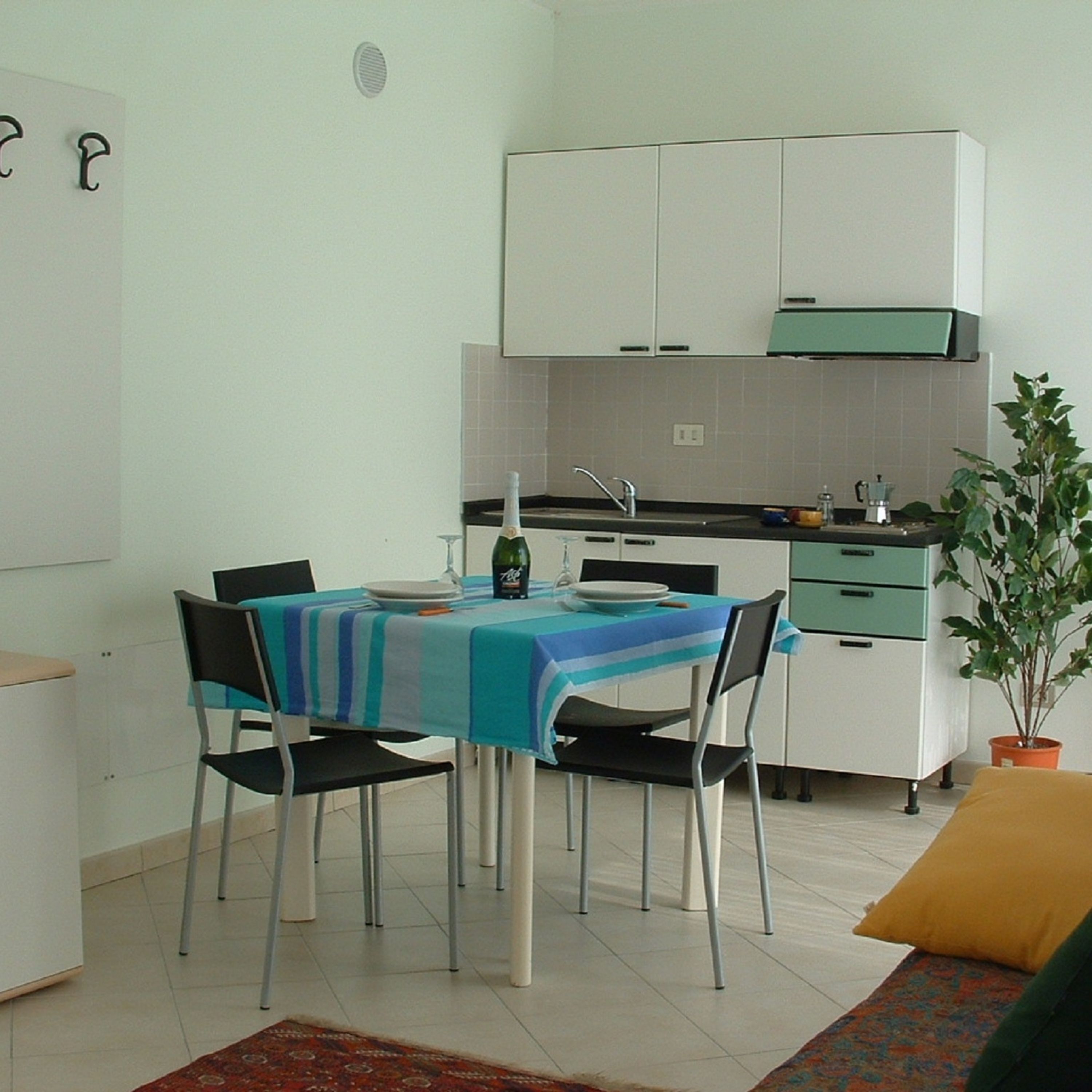 A small kitchen with white cabinets and a green countertop. There is a dining table with a blue tablecloth and four chairs. A bottle of wine and a glass are on the table.
