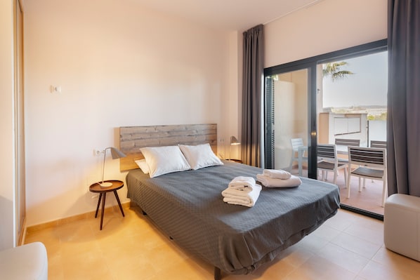 Une chambre d’hôtel moderne avec un grand lit, une petite table de chevet, une lampe et une vue sur l’espace extérieur aménagé en salon.