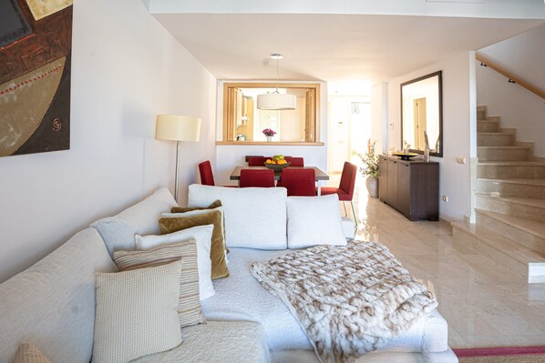 A modern living room with a sofa, a dining table with chairs, a staircase, and a large mirror.