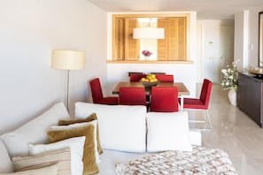 A modern living room with a sofa, red chairs, a dining table with a fruit bowl, and a lamp.