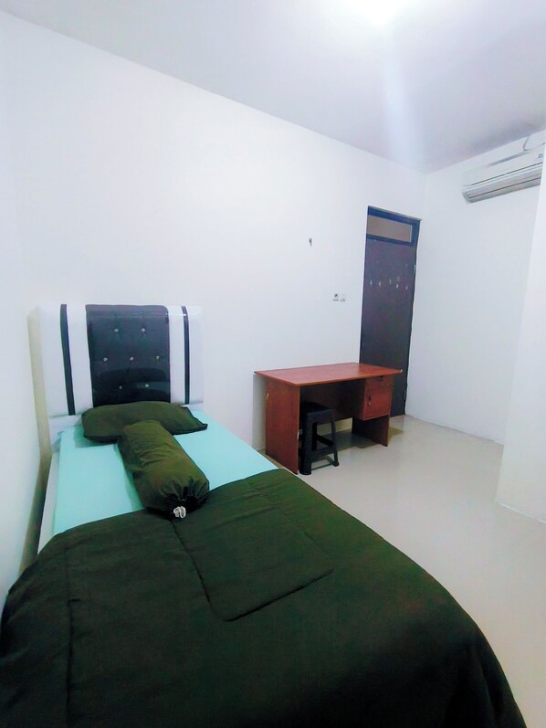 Una cama individual con una colcha verde y dos almohadas, un escritorio de madera y una puerta cerrada.