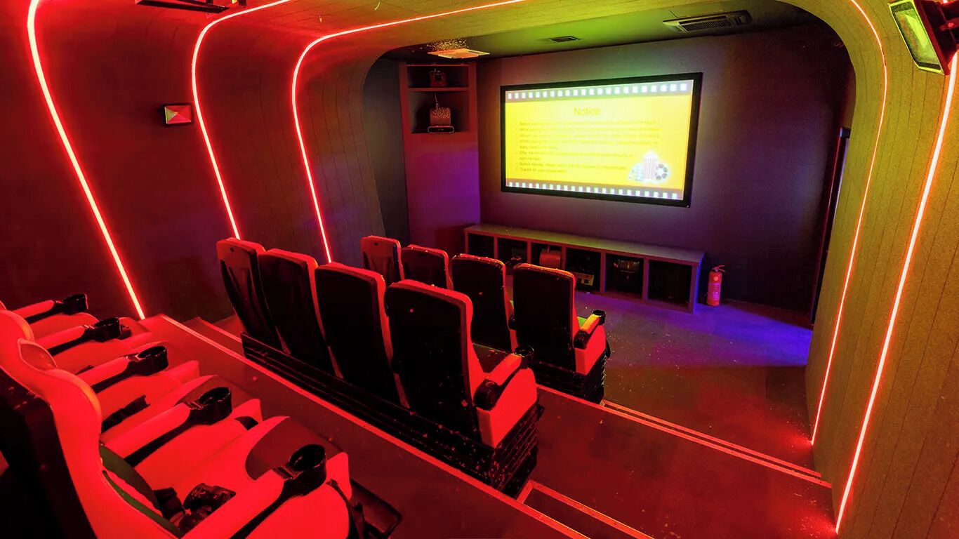 Une salle de cinéma éclairée par des néons rouges, avec des rangées de sièges, un écran affichant un avis et une petite scène.