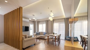 Een moderne woonkamer met een bank, salontafel, eettafel en stoelen. Links is een houten scheidingswand, aan de muur een flatscreen televisie en grote ramen met gordijnen.