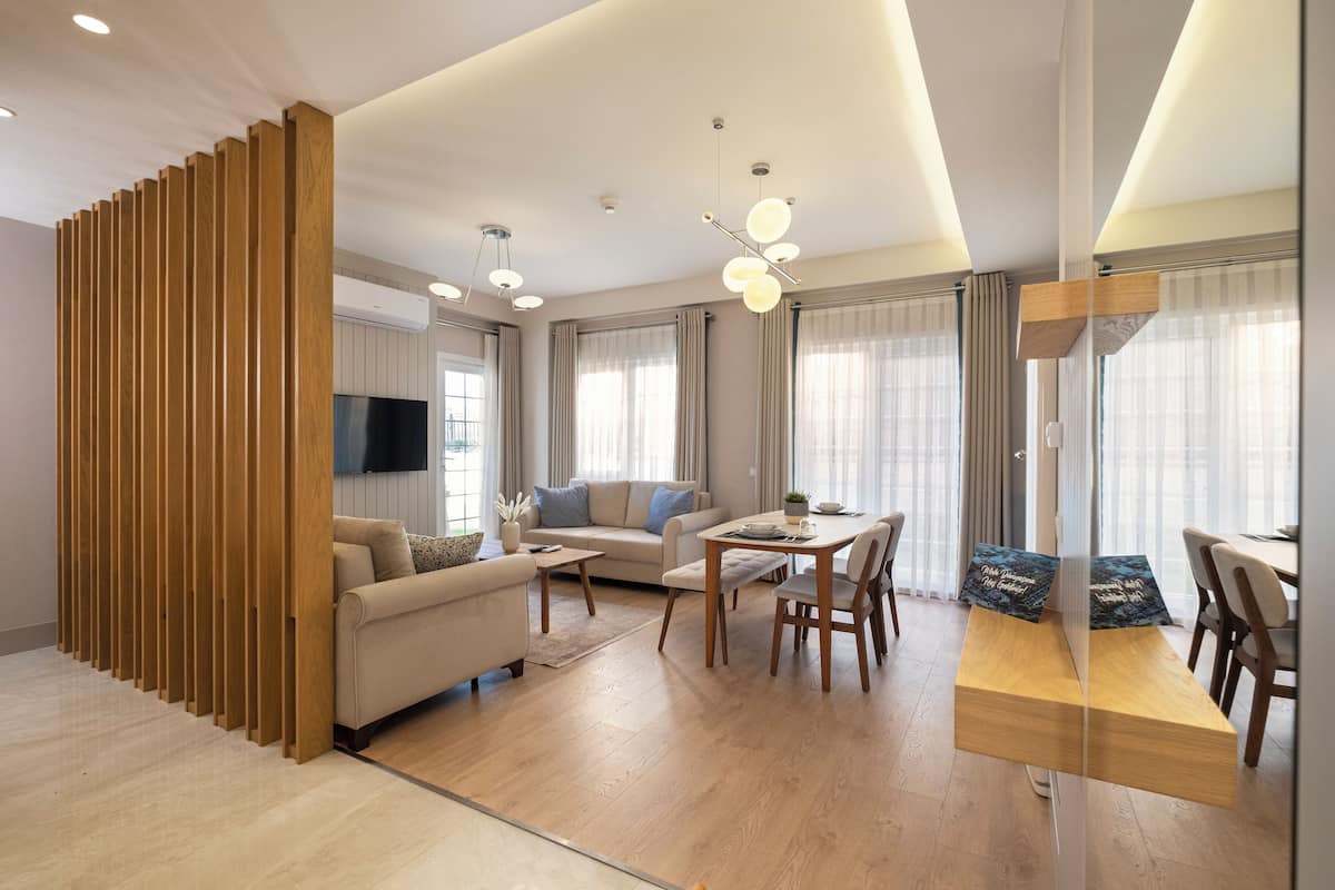 A modern living room with a sofa, coffee table, dining table, and chairs. There is a wooden partition on the left, a flat-screen TV mounted on the wall, and large windows with curtains.