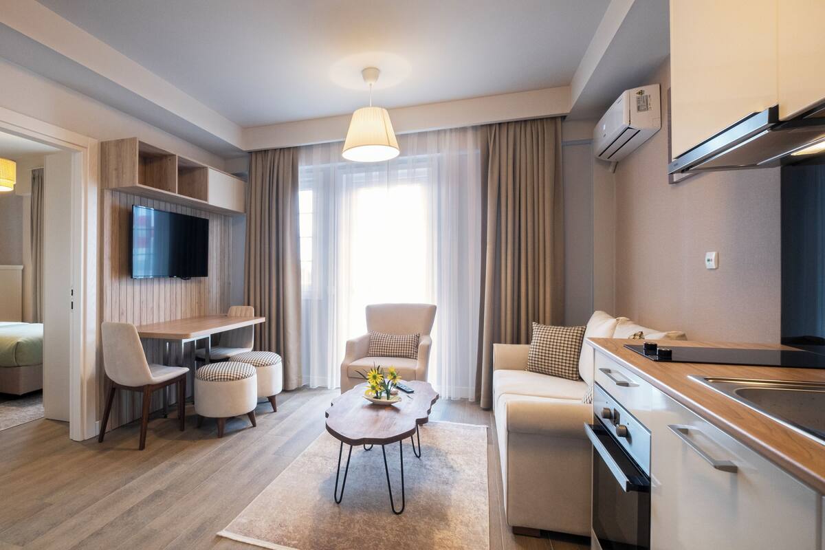 A modern hotel room with a dining area, a sofa, a TV mounted on the wall, and a kitchen area.
