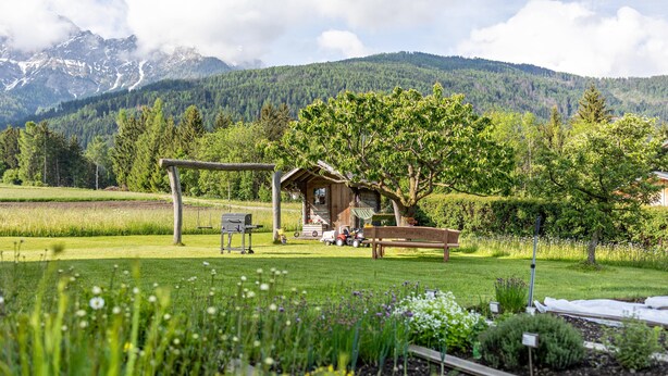 Una cabaña rústica con porche de madera, rodeada por un jardín cuidado y vistas a la montaña.