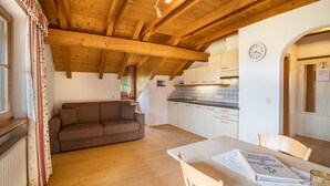 Un salon chaleureux avec un plafond en bois, un canapé marron, une table à manger avec un menu, et une cuisine en arrière-plan.