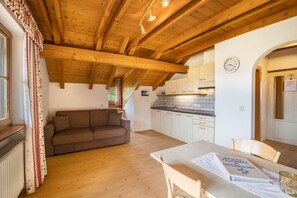Een gezellige zithoek met een houten plafond, een bruine bank, een eettafel met een menukaart, en een keuken op de achtergrond.