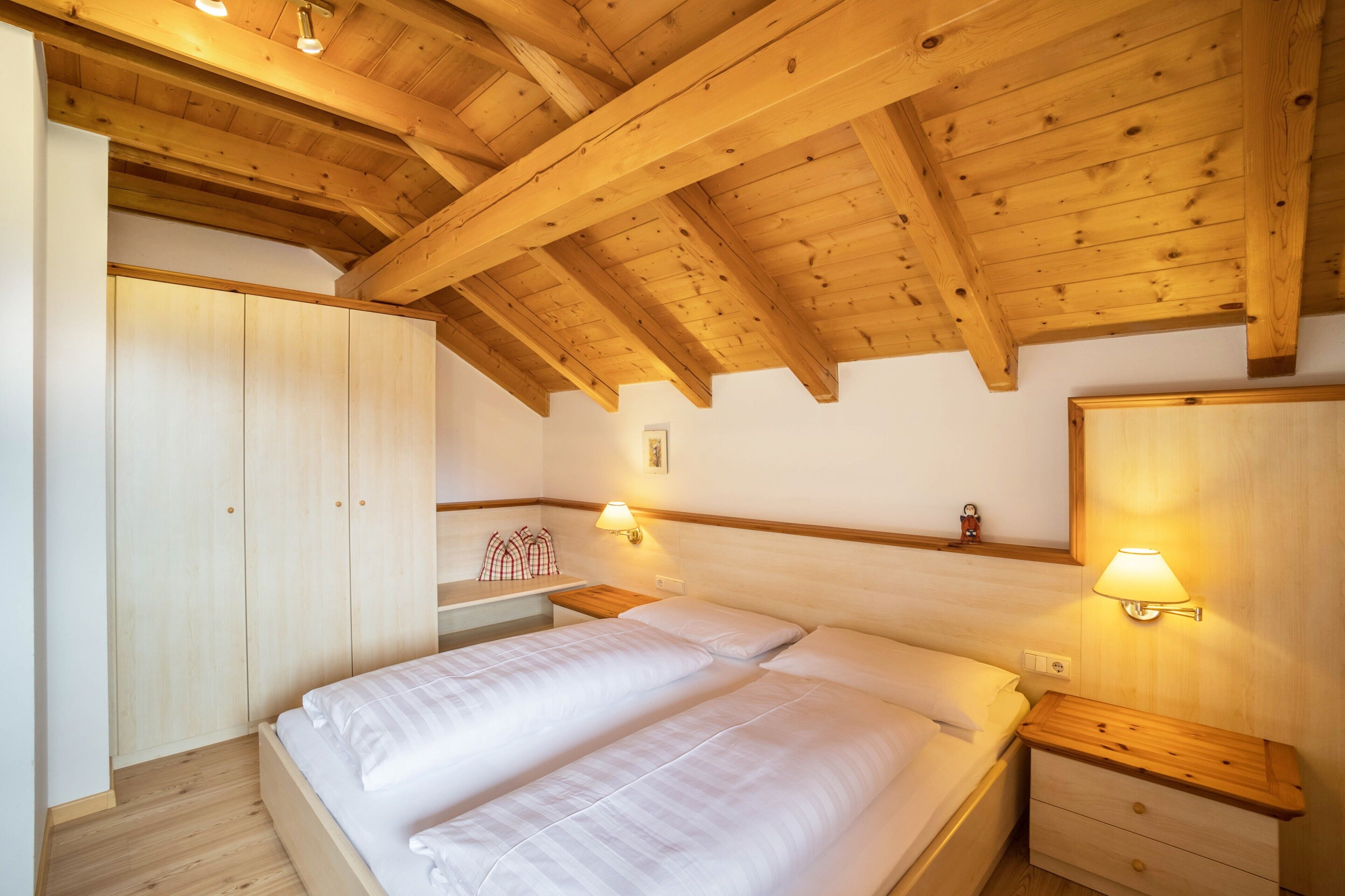 Un dormitorio con techo de madera, una cama con ropa de cama blanca, una mesita de noche, un armario y lámparas fijadas a la pared.